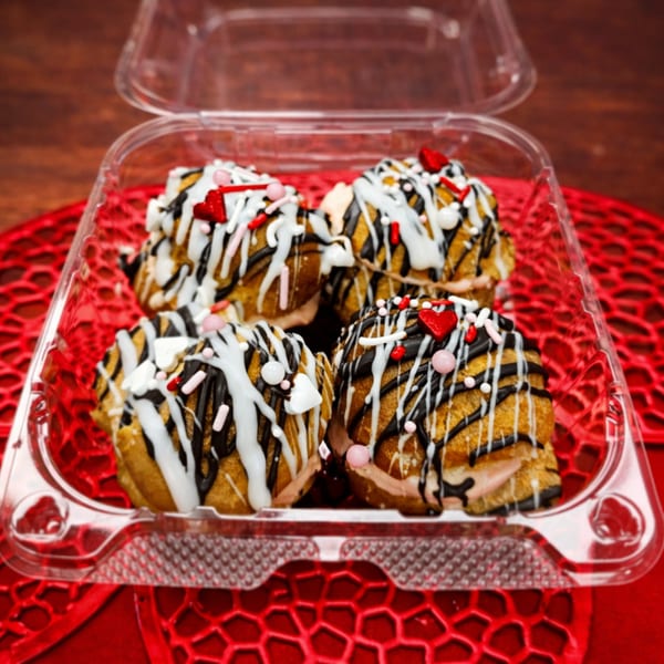 Four Italian cream puffs with chocolate and white icing, sprinkles, and heart candies in a plastic container.